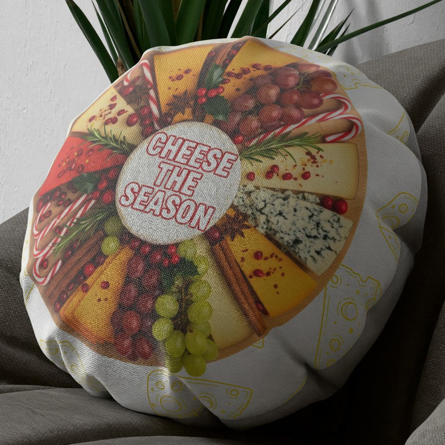 A round decorative throw pillow featuring a festive cheese board print with the words 'CHEESE THE SEASON' in the center, resting on a gray couch with a green plant in the background.