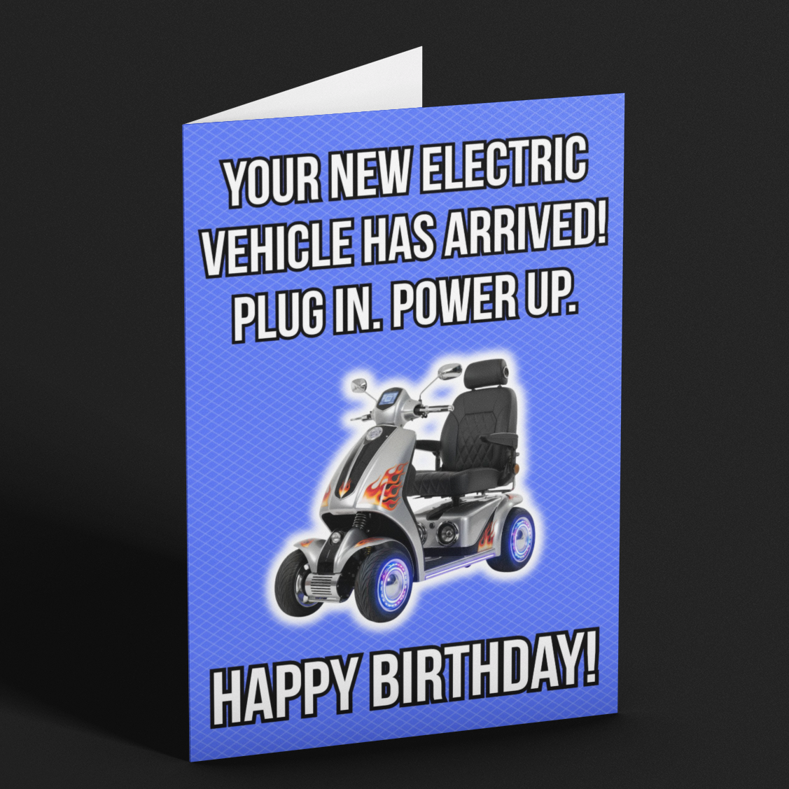 A folded birthday card standing upright on a dark background. The card has a bright blue background with white text reading 'YOUR NEW ELECTRIC VEHICLE HAS ARRIVED! PLUG IN.