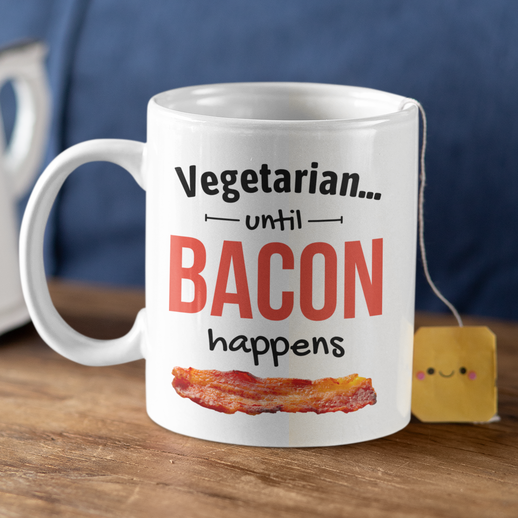 Mug with 'Vegetarian... until Bacon happens' text on a wooden surface with a teapot and tea bag.