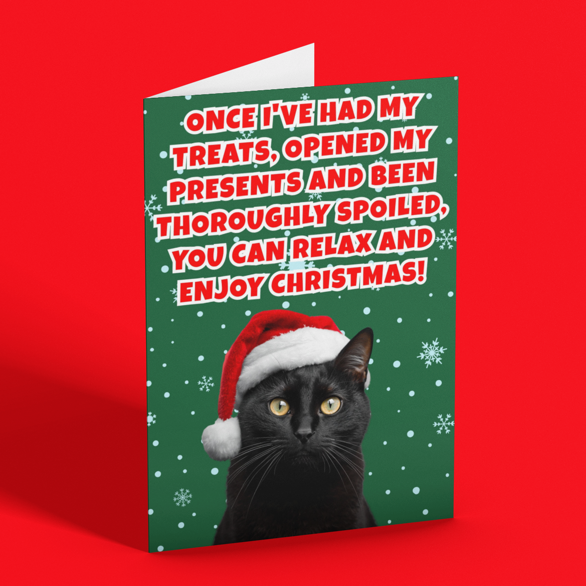 A Christmas greeting card featuring a black cat in a Santa hat on a green background with white snowflakes and red text, standing upright on a bright red surface.