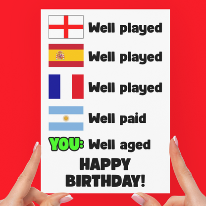A white greeting card held up by two hands against a solid red background. The card features rows of national flags with the text 'Well played' and 'Well paid', with the final line highlighted in green and black as 'YOU: Well aged HAPPY BIRTHDAY!'.
