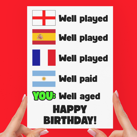 A white greeting card held up by two hands against a solid red background. The card features rows of national flags with the text 'Well played' and 'Well paid', with the final line highlighted in green and black as 'YOU: Well aged HAPPY BIRTHDAY!'.