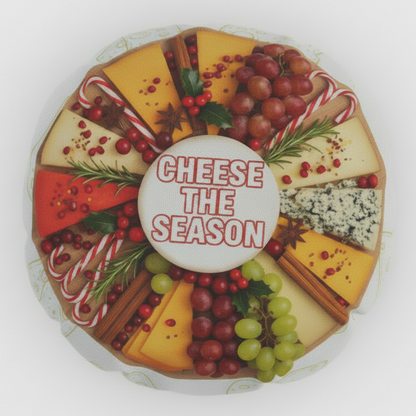 A round cheese board or cheese wheel with multiple cheese wedges arranged in a circle, decorated with red cranberries, green and purple grapes, cinnamon sticks, rosemary, and candy canes, with a white center label reading 'CHEESE THE SEASON'.