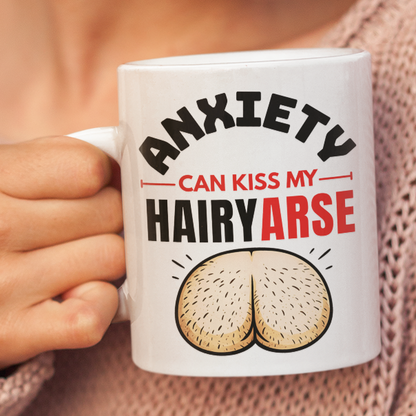 A woman in a pink knit sweater holds a white ceramic mug with the text 'ANXIETY CAN KISS MY HAIRYARSE' and a graphic of two buttocks. The background is a solid, warm beige color.