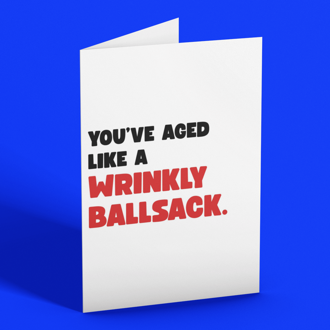 A white greeting card standing upright on a bright blue background, printed with the text 'YOU'VE AGED LIKE A WRINKLY BALLSACK.' in black and red lettering.