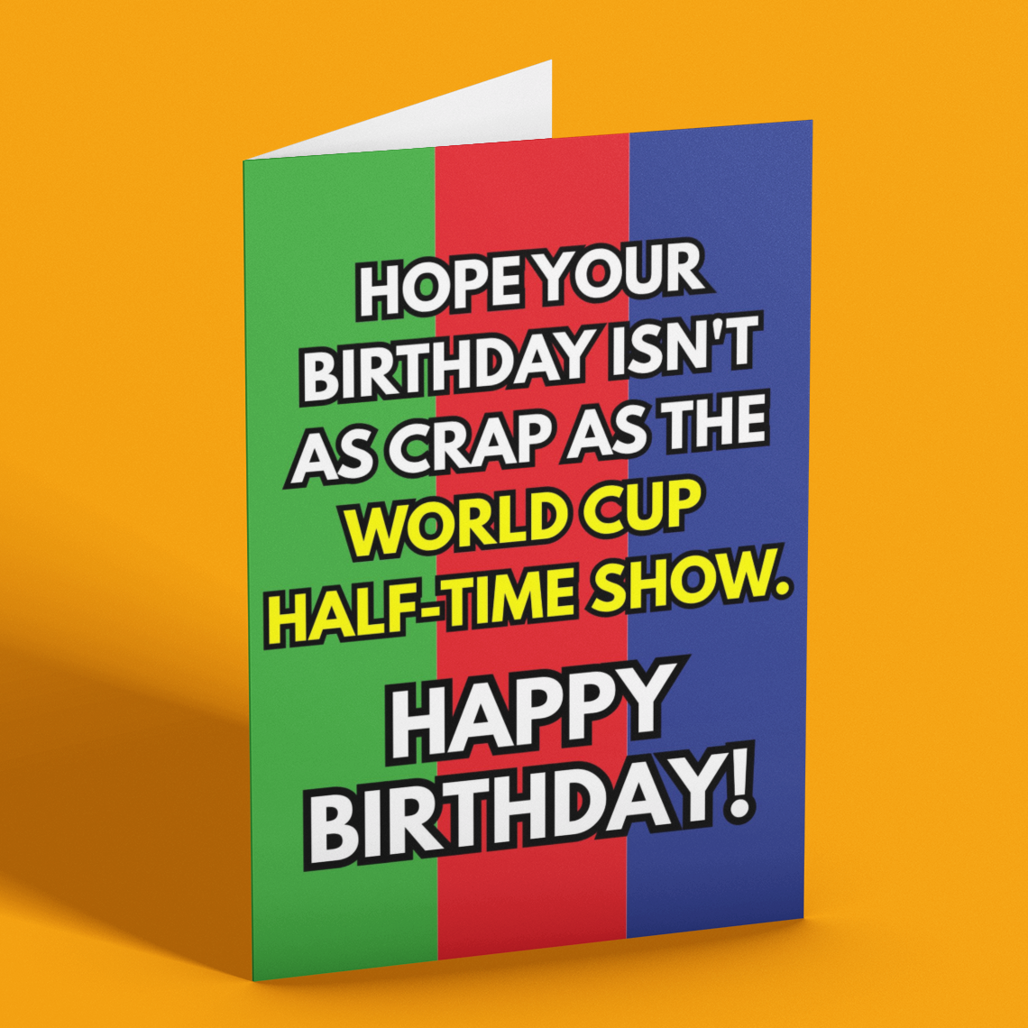 A folded greeting card standing upright on a solid orange background. The card features a colorful design with vertical stripes of green, red, and blue, and bold white text with black outlines that reads, 'HOPE YOUR BIRTHDAY ISN'T AS CRAP AS THE WORLD CUP HALF-TIME SHOW.