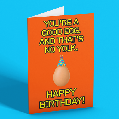 A folded greeting card standing upright on a bright blue background. The card is orange with yellow text reading 'YOU'RE A GOOD EGG.