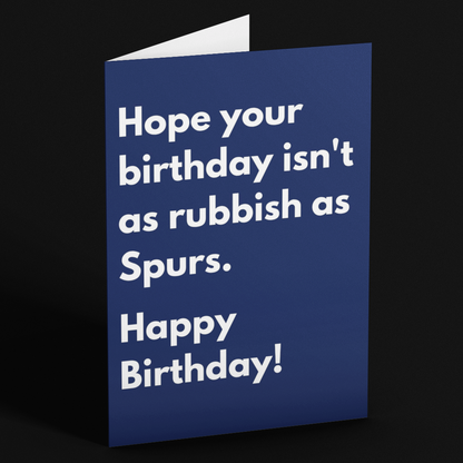 A folded blue greeting card standing upright on a black background. The front of the card has white text that reads, 'Hope your birthday isn't as rubbish as Spurs.