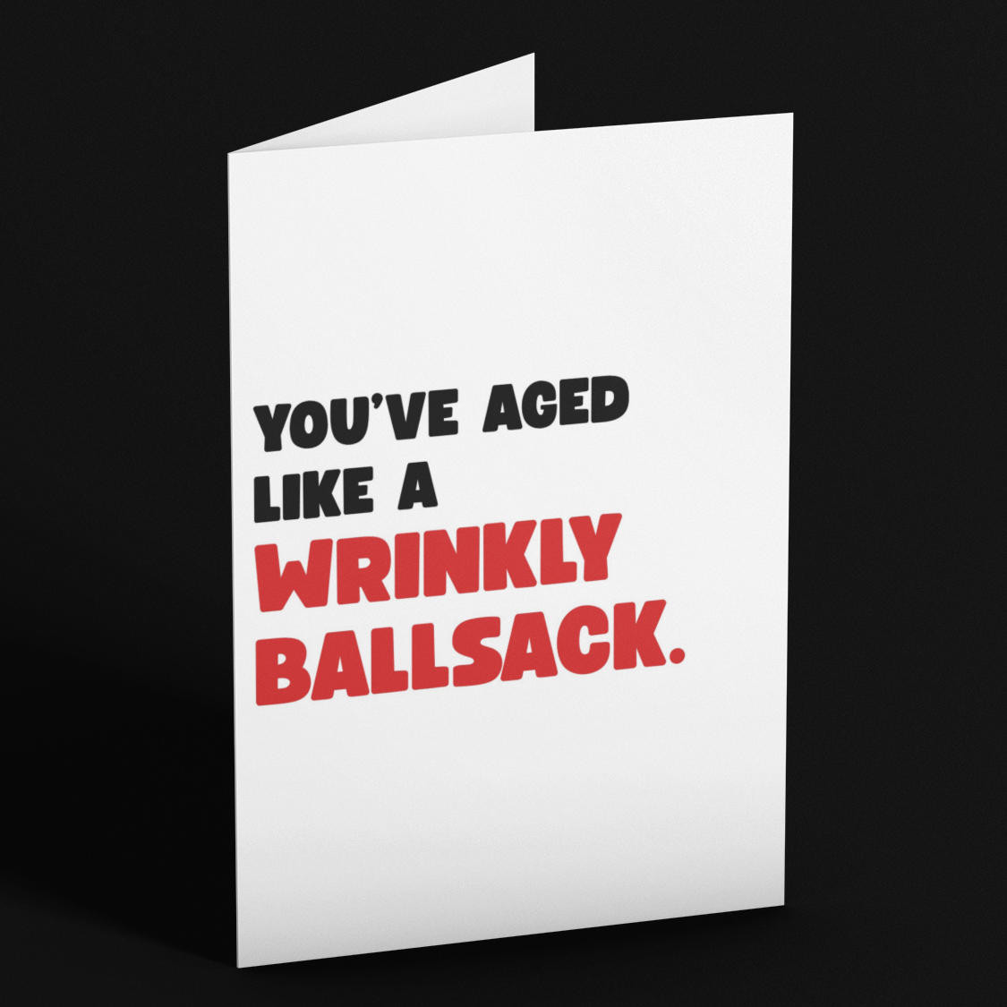 A white greeting card standing upright on a black background, printed with the text 'YOU'VE AGED LIKE A WRINKLY BALLSACK.' in black and red lettering.