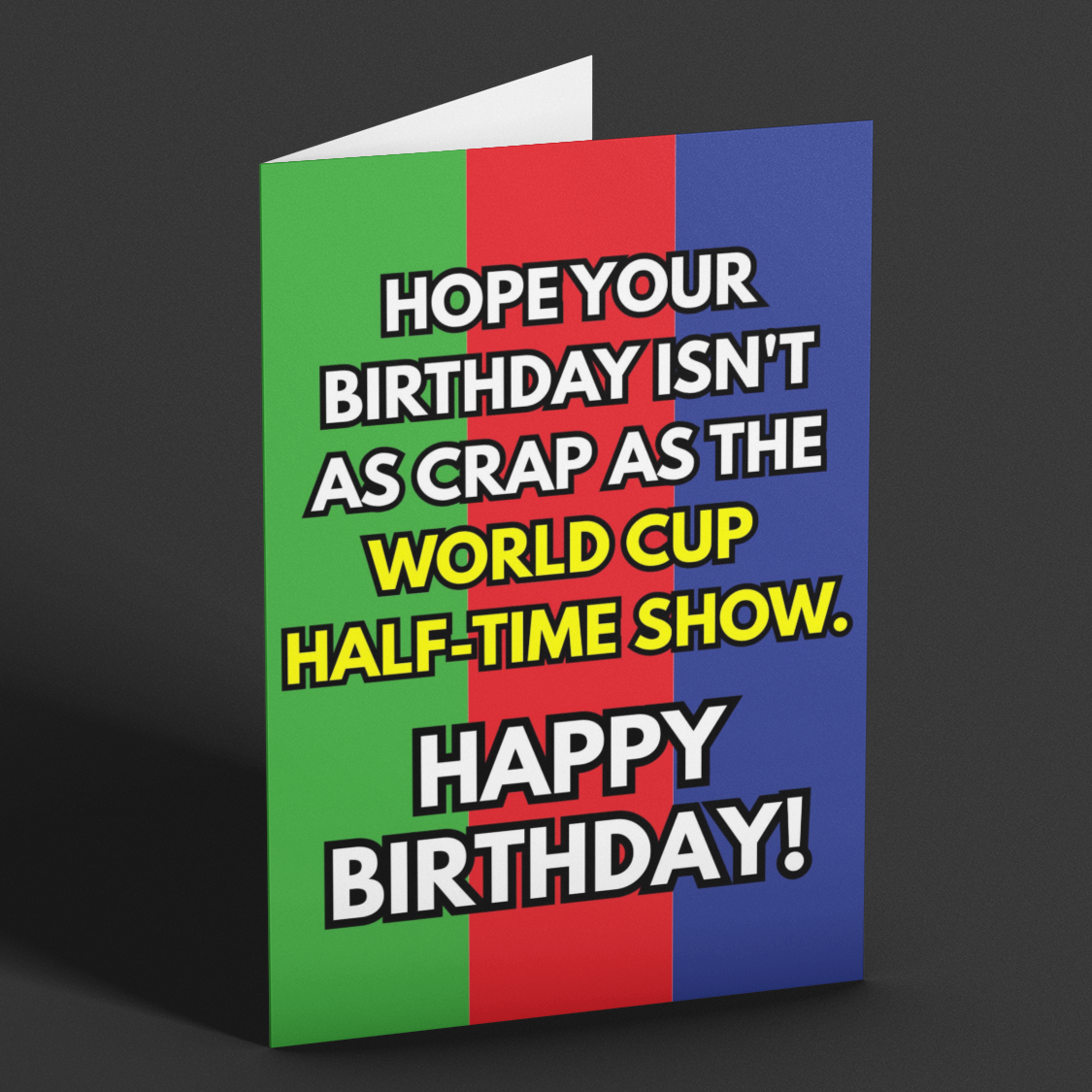 A folded greeting card standing upright on a dark gray background. The card has a bold design with vertical green, red, and blue panels and large white text with black outlines reading, 'HOPE YOUR BIRTHDAY ISN'T AS CRAP AS THE WORLD CUP HALF-TIME SHOW.
