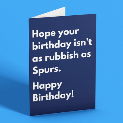 A navy blue greeting card with white text reading 'Hope your birthday isn't as rubbish as Spurs. Happy Birthday!' standing upright on a bright blue background.