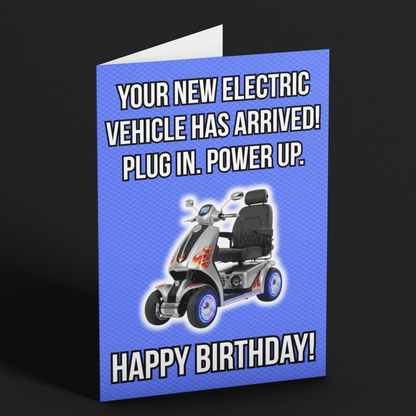 A folded birthday card standing upright on a dark background. The card has a bright blue background with white text reading 'YOUR NEW ELECTRIC VEHICLE HAS ARRIVED! PLUG IN.