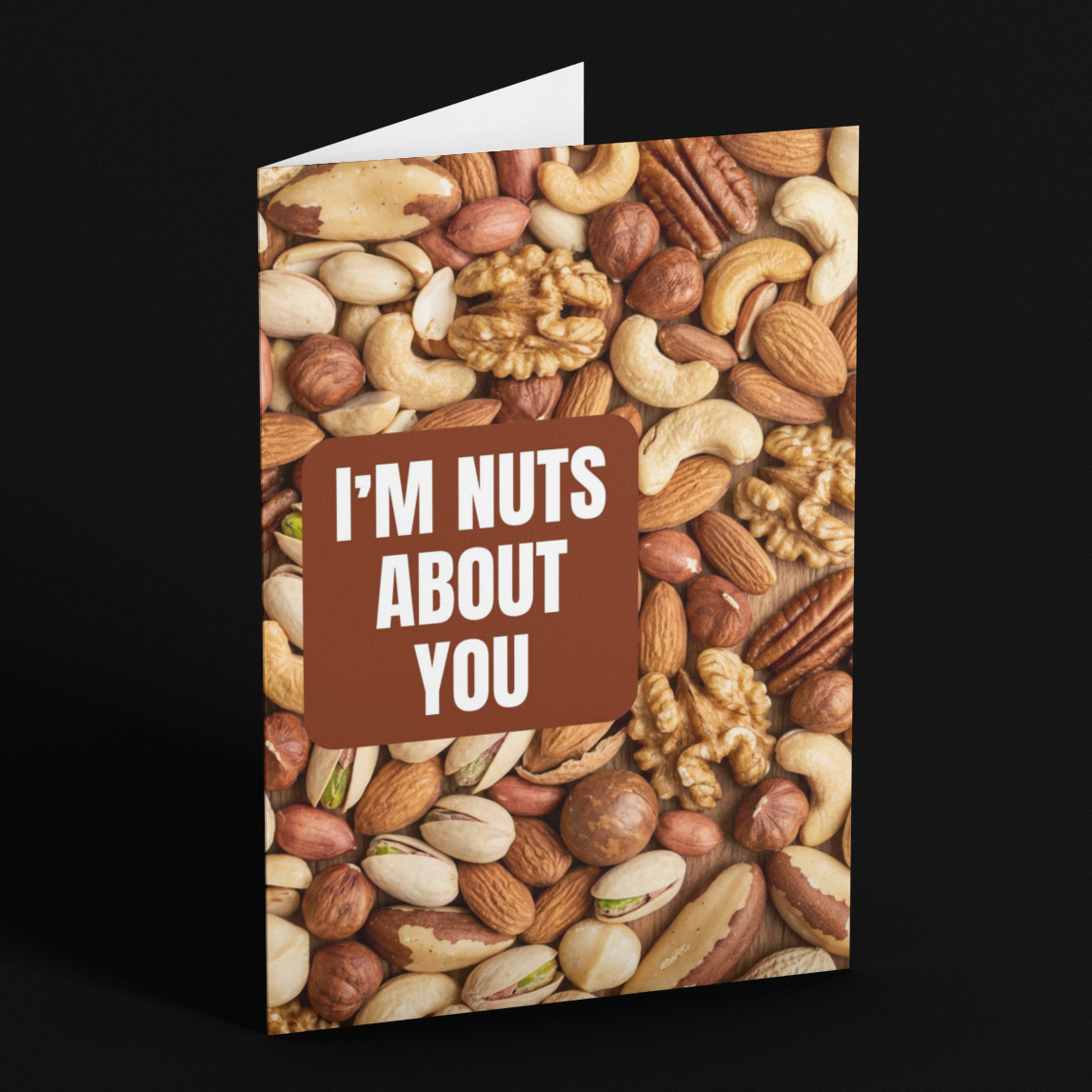 A folded greeting card standing upright on a black background. The card cover features a close-up photo of assorted nuts in tan, brown, cream, and beige tones, with a brown rectangular text panel in the center reading “I'M NUTS ABOUT YOU” in white capital letters.