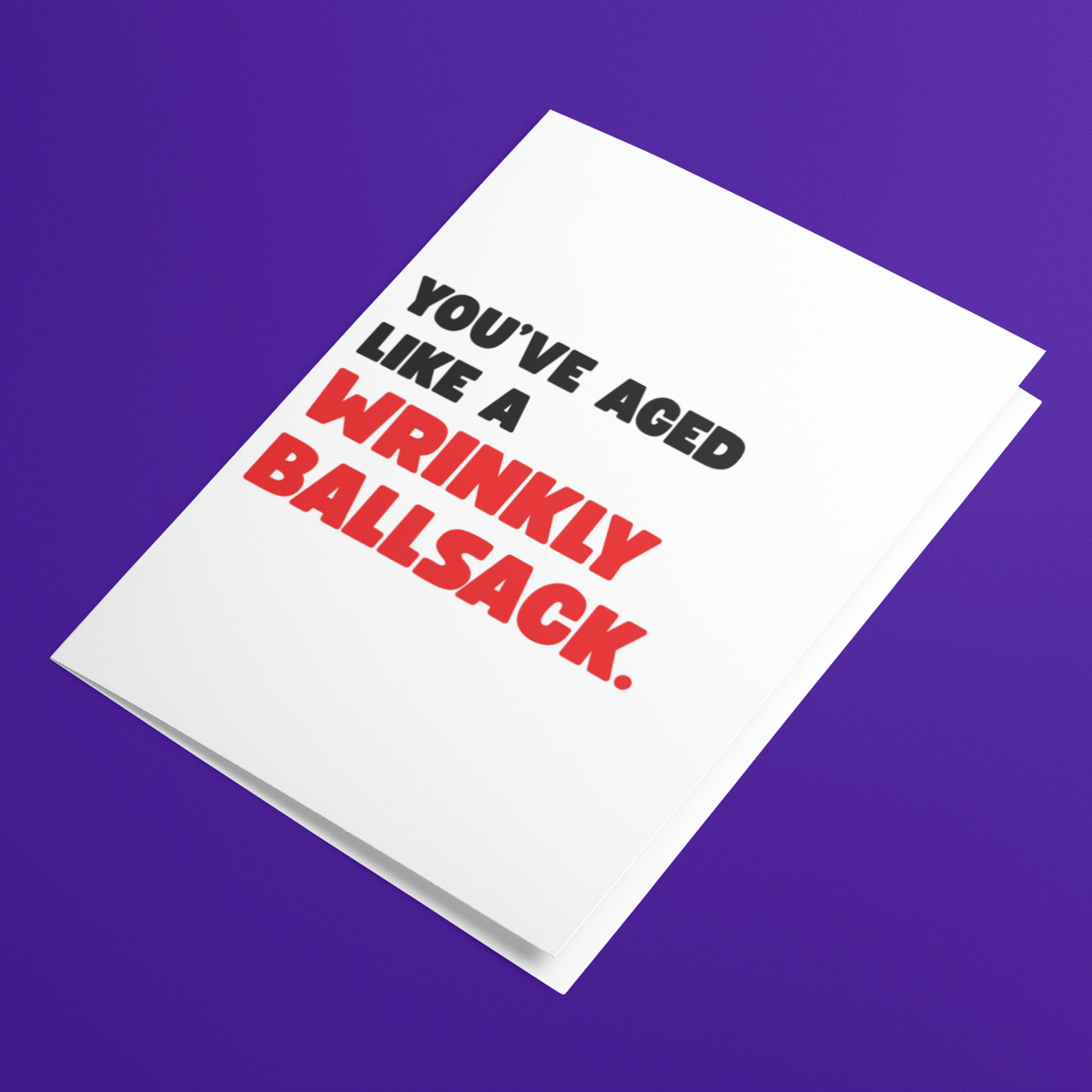 A white greeting card with bold black and red text reading 'YOU'VE AGED LIKE A WRINKLY BALLSACK.' shown at an angle on a solid purple background.