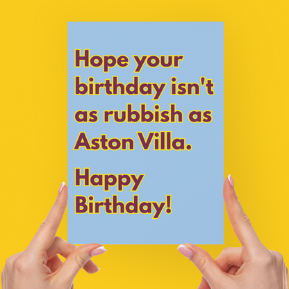 A person's hands holding a light blue greeting card with brown text that reads 'Hope your birthday isn't as rubbish as Aston Villa. Happy Birthday!' against a solid bright yellow background.