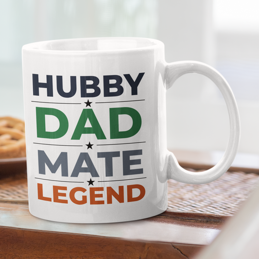 White mug with text 'HUBBY DAD MATE LEGEND' on a wooden table with pastries in the background