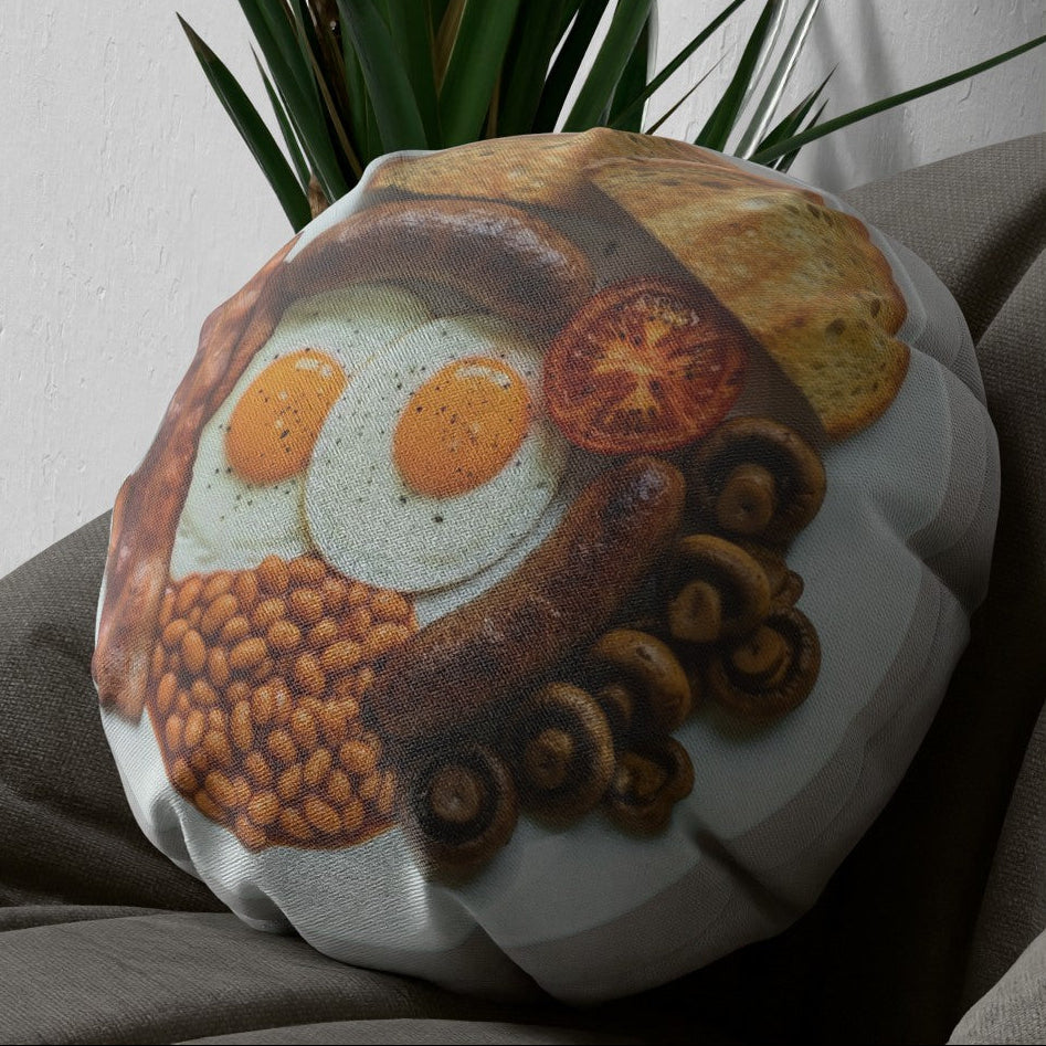 A round decorative throw pillow featuring a printed breakfast food illustration with eggs, sausage, beans, mushrooms, and toast, styled on a gray fabric surface with a green plant in the background.