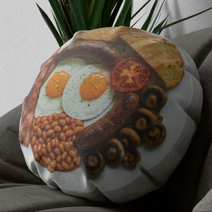 A round decorative throw pillow featuring a printed breakfast food illustration with eggs, sausage, beans, mushrooms, and toast, styled on a gray fabric surface with a green plant in the background.