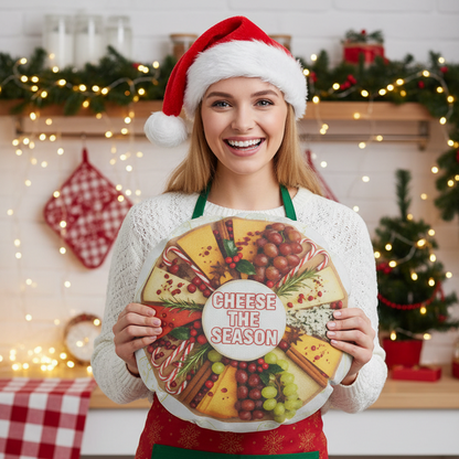 A smiling woman in a white sweater, red apron, and Santa hat holds a large round holiday sign that reads 'CHEESE THE SEASON' with a cheese wheel illustration. The background shows a decorated Christmas kitchen with warm lights, garlands, and wrapped gifts.