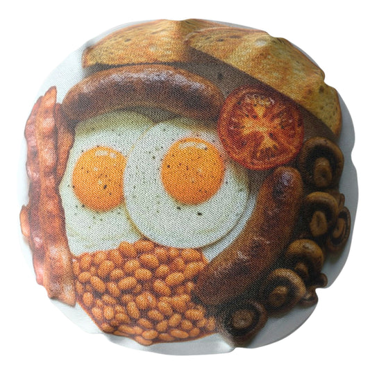 A round decorative pillow featuring a detailed print of a hearty breakfast food scene with two fried eggs, a sausage, bacon, baked beans, toast, and a tomato slice on a white background.
