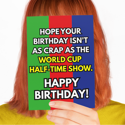 A person with red hair wearing a yellow sweatshirt holds a colorful birthday card in front of their face. The card has a green, red, and blue background with bold white text that reads, 'HOPE YOUR BIRTHDAY ISN'T AS CRAP AS THE WORLD CUP HALF-TIME SHOW.